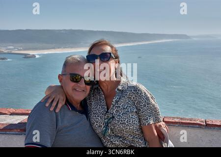 Ein älteres Ehepaar posiert zärtlich umarmt an einem Aussichtspunkt in Nazare, Portugal, mit dem Atlantik und der Küste im Hintergrund. Stockfoto
