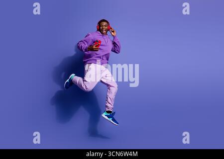 Ein energiegeladener junger Mann in lila Mode springt mit Telefon und Kopfhörern und zeigt einen stilvollen, lässigen Lifestyle Stockfoto