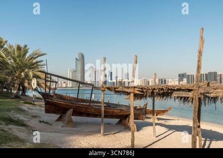 Vereinigte Arabische Emirate - Abu Dhabi - Heritage Village - Dau am Strand, Palmenschatten und Skyline über das ruhige Wasser des Golfs erinnern an den dauerhaften maritim der Stadt Stockfoto