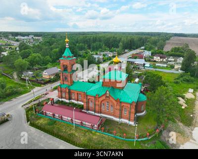 Ein Blick aus der Vogelperspektive auf die farbenfrohe Kirche aus rotem Backstein in Petushki, die sich auf einer langen, geraden Straße befindet, umgeben von grünen Feldern und Wäldern. Stockfoto