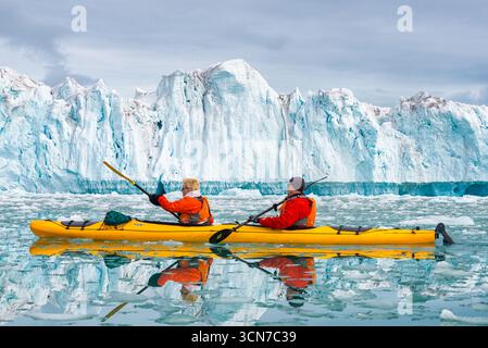 Kajakfahrer paddeln in Klerckbukta Nordaustlandet Svalbard Norwegen // NORDEAUSTLANDET, SVALBARD UND JAN MAYEN — zwei Kajakfahrer paddeln durch eiskalte arktische Gewässer vor einer riesigen Gletscherwand in Klerckbukta. Die leuchtend gelben Kajaks und orangen Schwimmwesten der Paddler stehen in starkem Kontrast zu den eisblauen und weißen Tönen des Gletschers und des umliegenden Meereises. Klerckbukta liegt an der nordöstlichen Küste von Nordaustlandet, der zweitgrößten Insel im Svalbard-Archipel. Diese abgelegene Region ist bekannt für ihre dramatischen Gletscherlandschaften und ist ein beliebtes Ziel für arktische Expeditionen Stockfoto