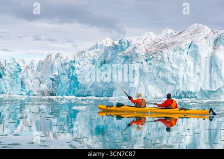 Kajakfahrer paddeln vorbei an der Bråsvellbreen Gletscherwand Nordaustlandet Svalbard Norwegen // NORDEAUSTLANDET, Svalbard — zwei Kajakfahrer in knallgelben Kajaks navigieren durch die eisigen Gewässer in der Nähe von Nordaustlandet, Svalbard, Norwegen, mit einer riesigen Gletscherwand als Kulisse. Die lebendige Farbe der Kajaks steht im Kontrast zu den tiefen Blau- und Weißtönen des Gletschereises und des umliegenden Meereises. Nordaustlandet ist die zweitgrößte Insel im Svalbard-Archipel im Arktischen Ozean. Es ist bekannt für seine ausgedehnten Eiskappen und dramatischen Gletscherlandschaften, was es zu einem erstklassigen Reiseziel für arktische Expedit macht Stockfoto