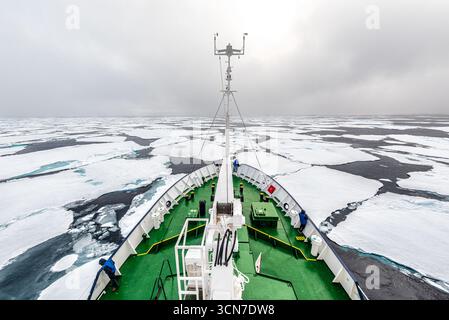 Ship Bow in Arctic Sea Ice Svalbard Norwegen / SVALBARD UND JAN MAYEN — der Bug eines Expeditionsschiffs schiebt durch arktisches Meereis in den Gewässern bei Sjuøyane, dem Sieben-Inseln-Archipel im Norden von Svalbard. Mehrere Figuren in blauen Jacken stehen auf dem grün gestrichenen Deck und beobachten die zerbrochenen Schollen, die sich um den Rumpf vor einem bewölkten Polarhimmel drehen. Die Navigation auf dem Packeis um Sjuøyane erfordert eisverstärkte Schiffe oder Eisbrecher; die gleichen Gewässer hielten HMS Racehorse und HMS Carcass während der Phipps-Expedition 1773 ein, was die Besatzungen dazu zwang, ihre Schiffe und hau zu verlassen Stockfoto