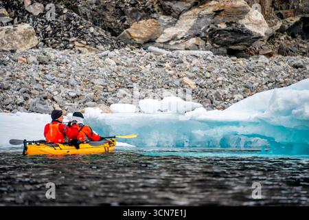Kajakfahrer in Orange Life Jackets Paddeln Sie vorbei an Blue Iceberg Svalbard Norwegen / SVALBARD UND JAN MAYEN — zwei Kajakfahrer in leuchtend orange Schwimmwesten paddeln mit einem gelben Kajak an einem leuchtend blauen Eisberg in den arktischen Gewässern um Sjuøyane, den Sieben Inseln Archipel im Norden von Svalbard. Die zerklüftete, felsige Küste der Inseln bildet eine schroffe Kulisse für das Eis, das im dunklen Wasser schwimmt. Sjuøyane ist das nördlichste Land Norwegens und Teil des Naturreservats Nordaust-Svalbard, wodurch Kajakexpeditionen hier zu den abgelegensten arktischen Paddelerlebnissen gehören. Stockfoto