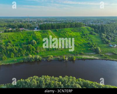 Ein Panoramablick auf einen breiten, geschwungenen Fluss, der an einem grünen, grasbewachsenen Hügel mit Wanderwegen vorbei fließt, mit einem ländlichen Dorf und Wald im Hinterland Stockfoto