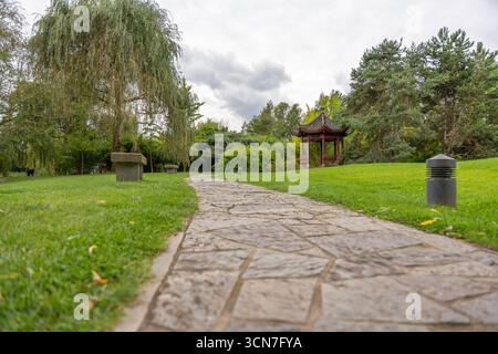 Ein Steinweg führt durch einen Park mit Bänken und einem traditionellen chinesischen Pavillon. Üppige Bäume und Gras umgeben den Gehweg. Stockfoto