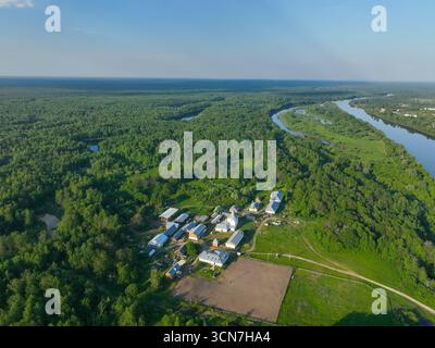 Eine weite Luftaufnahme eines Bauernhofs mit Gebäuden und Feldern, neben einem riesigen grünen Wald und einem sich windenden Fluss unter einem klaren blauen Himmel. Stockfoto