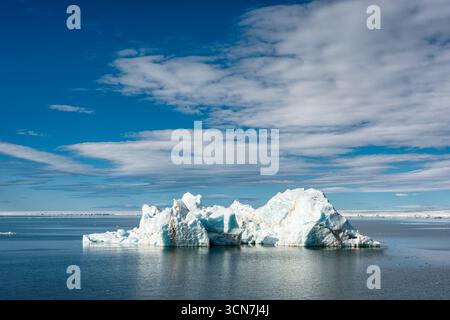 Eisberg in Klerckbukta Nordaustlandet Svalbard Norwegen // KLERCKBUKTA, Nordaustlandet, Svalbard — Ein großer Eisberg driftet in den arktischen Gewässern von Klerckbukta, einer Bucht auf Nordaustlandet, der zweitgrößten Insel im Svalbard-Archipel. Der Eisberg weist komplizierte Formationen und verschiedene weiß- und Blautöne auf, die charakteristisch für glaziales Eis sind. Im Hintergrund ist eine ferne Eiskante oder Küste unter einem hellblauen Himmel mit weißen Wolken zu sehen. Nordaustlandet ist Teil des Svalbard and Jan Mayen Territory, einer Inselgruppe im Arktischen Ozean. Diese Region ist bekannt für Stockfoto