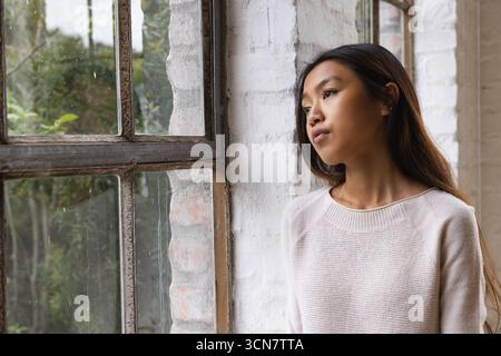 Asiatische Frau, die am Fenster steht, mit Regentropfen in weißem Ziegelloft, die nach draußen schauen, Kopierraum Stockfoto