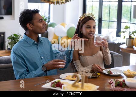 Indisches Paar toast Geburtstag mit Gläsern am heimischen Tisch neben Kuchen, Mädchen im Diadem, Kopierraum Stockfoto