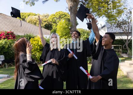 Verschiedene Klassenkameraden auf dem Rasen in schwarzen Kleidern, die Mortarboards mit Diplomen und Bändern werfen Stockfoto