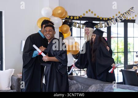 Verschiedene Absolventen in Kleidern, die sich umarmen und Diplome halten, Ballons im Wohnzimmer, Kopierraum Stockfoto