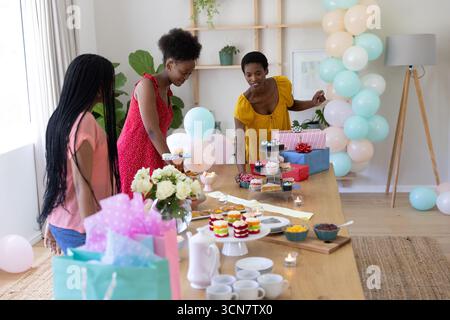 Afroamerikanische Freunde bereiten Cupcakes auf dem Tisch unter pastellfarbenen Ballons im hellen Speisesaal zu Stockfoto