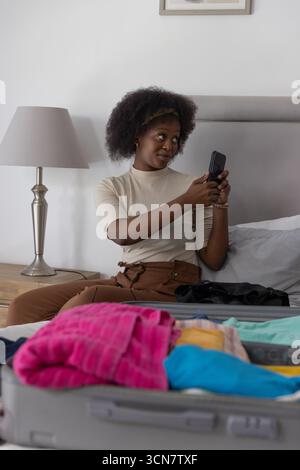 Afroamerikanerin packt Kleidung in den Koffer und hält das Smartphone, während sie auf dem Bett sitzt Stockfoto