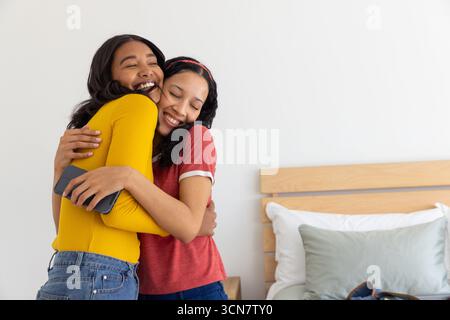 Verschiedene Mutter und Teenager-Tochter umarmen sich im Schlafzimmer und halten das Smartphone zwischen Kissen auf dem Bettgestell Stockfoto