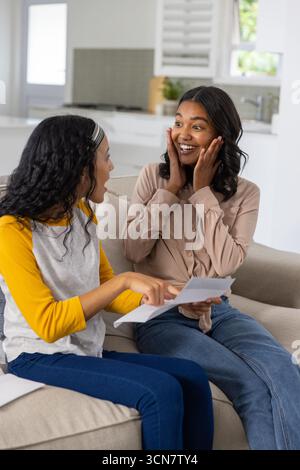 Verschiedene Mom und Teenager-Tochter halten Papierlaken und sitzen auf beigefarbenem Sofa neben Küchenspüle Stockfoto
