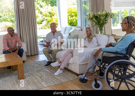 Verschiedene Seniorenfreunde unterhalten sich um einen Couchtisch im Wohnzimmer mit Rollstuhl Stockfoto