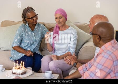 Verschiedene ältere Freunde sitzen auf der Couch im Wohnzimmer und feiern mit Kuchen mit zündeten Kerzen, Tassen Stockfoto