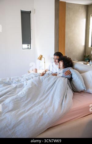 Asiatischer amerikanischer Mann und Afroamerikaner kuscheln sich unter grauer Decke im Schlafzimmer mit Messinglampe Stockfoto