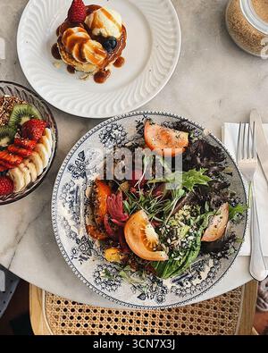 Gourmet Cafe Brunch Aufstrich: Quinoa-Salat, Pfannkuchen mit Karamellsauce und eine gesunde Müsli- und Obstschale mit Kiwi, Erdbeeren und Banane. Stockfoto