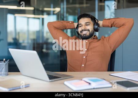 Junger indischer Mann, der Kopfhörer trägt und lächelt, eine Pause von der Arbeit im Büro macht, sich mit Händen hinter dem Kopf zurück im Stuhl lehnt und einen Moment der Entspannung genießt Stockfoto