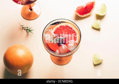 Cocktailglas mit Grapefruitscheiben, Eiswürfeln und Rosmarin auf beigefarbenem Hintergrund. Erfrischendes Sommergetränk, Blick von oben. Stockfoto