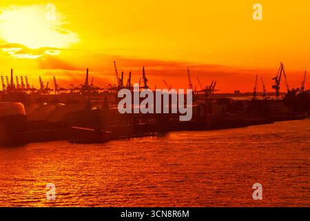 Der Sonnenuntergang taucht einen belebten Frachthafen in ein goldenes Leuchten, die Kräne vor dem dramatischen Himmel umrahmen und sich intensiv auf dem Wasser spiegeln. Fesselnde Szene Stockfoto