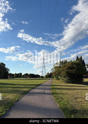 Stromturm mit Hochspannungsleitungen, die sich über einen hellblauen Himmel mit weißen Wolken erstrecken. Ein gepflasterter Pfad führt durch ein breites grünes Feld W Stockfoto