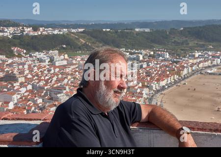 Ein Mann mittleren Alters mit einem markanten weißen Bart und einem freundlichen Lächeln lehnt sich an eine Wand und blickt auf die Landschaft. Hinter ihm erstreckt sich ein atemberaubendes Panorama Stockfoto