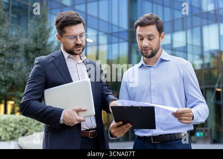 Zwei professionelle Geschäftsleute arbeiten an einem neuen Projekt zusammen, prüfen wichtige Dokumente und verwenden einen Laptop, während sie außerhalb eines modernen Bürogebäudes stehen Stockfoto