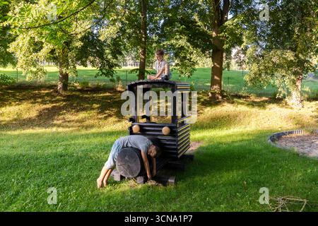 Zwei Kinder spielen glücklich auf einem hölzernen Eisenbahnbau in einem leuchtend grünen Park. Die Sonne scheint, und die Natur umgibt sie und schafft eine Freude Stockfoto