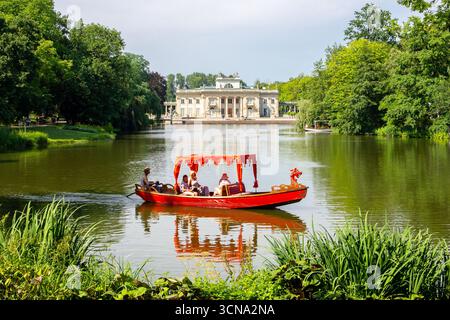 Warschau, Polen, 13.07.21. Rote Gondel mit Touristen auf dem See mit dem Palast auf der Insel (Polnisch: Palac Na Wyspie) im Hintergrund. Stockfoto