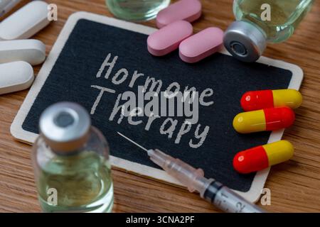 Verschiedene Pillen, Kapseln und Durchstechflaschen mit einer Spritze auf einem Holztisch. Eine kleine Tafel in der Mitte trägt die Worte „Hormontherapie“. Stockfoto