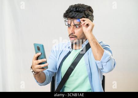 Ein junger Mann mit schockiertem Ausdruck, der seine Brille hält, während er auf sein Handy blickt. Mann überrascht, als er das Telefon ansah und Brillen anstellte. Stockfoto