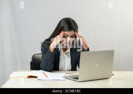 Titel: Eine frustrierte junge Geschäftsfrau mit dem Kopf in den Händen, die mit einem Laptop an einem Schreibtisch sitzt. Stockfoto