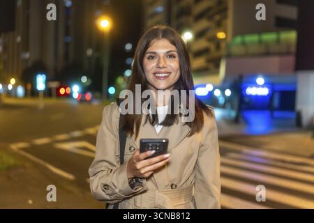 Frau lächelt hell und hält ein Smartphone, steht nachts auf einer belebten Stadtstraße mit beleuchteten Gebäuden und Verkehrsampeln, die einen erzeugen Stockfoto