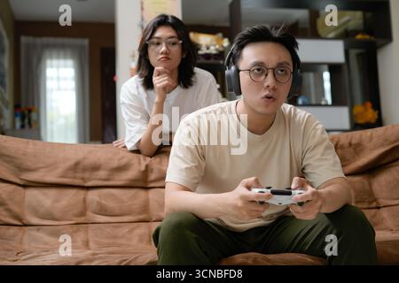 Ein Mann mit einem beigefarbenen Hemd sitzt auf einer braunen Couch und konzentriert sich auf einen Game-Controller mit Over-Ear-Kopfhörern, während eine Frau von hinten zusieht Stockfoto