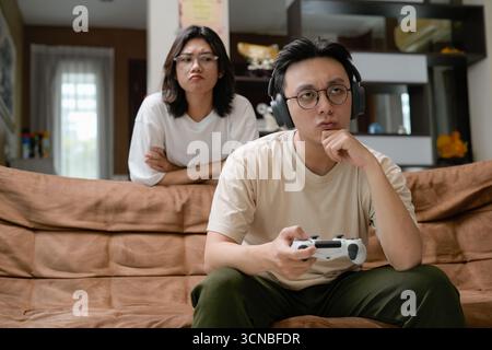Ein Mann mit einem beigefarbenen Hemd sitzt auf einer braunen Couch und konzentriert sich auf einen Game-Controller mit Over-Ear-Kopfhörern, während eine Frau von hinten zusieht Stockfoto