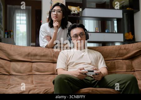 Ein Mann mit einem beigefarbenen Hemd sitzt auf einer braunen Couch und konzentriert sich auf einen Game-Controller mit Over-Ear-Kopfhörern, während eine Frau von hinten zusieht Stockfoto