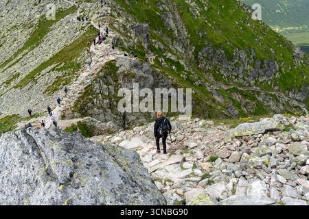 Entschlossene Wanderer mit Trekkingstöcken, die felsige Bergpfade in den Tatra-Gipfeln absteigen, mit mehreren Klettergruppen, die auf anspruchsvollen Alpin sichtbar sind Stockfoto