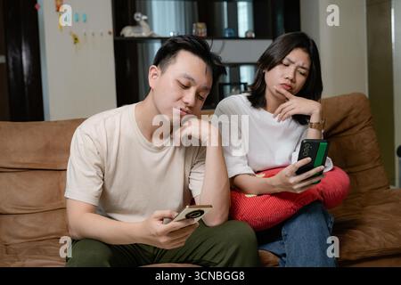 Zwangloses asiatisches Paar, das sich auf einer Couch in einem gemütlichen Wohnzimmer entspannt und auf die Telefone blickt Stockfoto