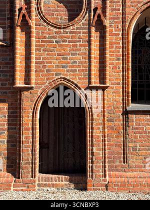 Nahaufnahme des Eingangs der gotischen Backsteinkirche aus dem Jahr 1370 in Europa, mit detaillierter, spitzbogenförmiger Holztür, die in roter Backsteinstruktur eingerahmt ist. Beispiel für Stockfoto