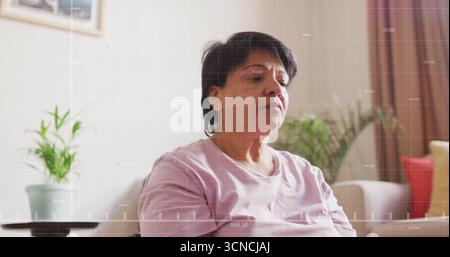 Sitzende indische Seniorin mit rosa T-Shirt, die im Wohnzimmer blickt, mit Pflanzen und rotem Kissen Stockfoto