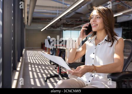 Sitzende Frau, die Dokumente in der Hand hält und auf dem Smartphone im Büro, im Kopierbereich spricht Stockfoto