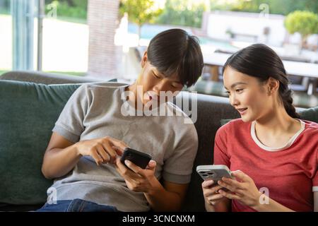 Asiatisches Paar, das auf dem Sofa sitzt und auf Smartphone-Bildschirme an der Tür mit Blick auf den Pool zeigt Stockfoto