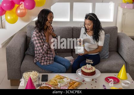 Vielfältige Mutter und Teenager-Tochter sitzen auf Wohnzimmer Sofa mit Geburtstagskuchen, bunten Ballons Stockfoto