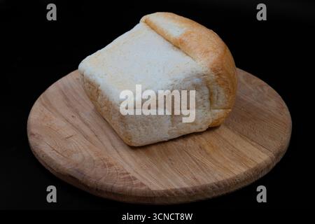 Frischer Brotlaib auf einem runden Holzbrett mit schwarzem Hintergrund. Perfekt für Bäckerei, hausgemachte Speisen, Frühstück, gesunde Ernährung und Culin Stockfoto