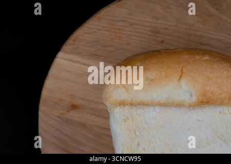 Frischer Brotlaib auf einem runden Holzbrett mit schwarzem Hintergrund. Perfekt für Bäckerei, hausgemachte Speisen, Frühstück, gesunde Ernährung und Culin Stockfoto