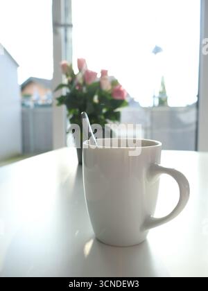 Eine helle, ruhige Nahaufnahme einer weißen Keramikbecher mit Kaffee auf einem sauberen Tisch, Löffel innen, mit einem verschwommenen rosa Bouquet im Hintergrund, fesselnd Stockfoto
