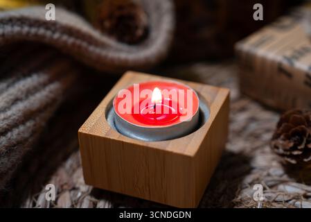 Minimalistische gemütliche Szene mit einer roten Kerze in einem quadratischen Holzhalter auf weicher Strickoberfläche und warmen Bokeh-Lichtern. Stockfoto